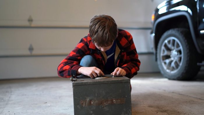 A young boy in a red plaid jacket squats on a concrete floor, intently manipulating an old metal box in a garage with a truck and a garage door in the background.