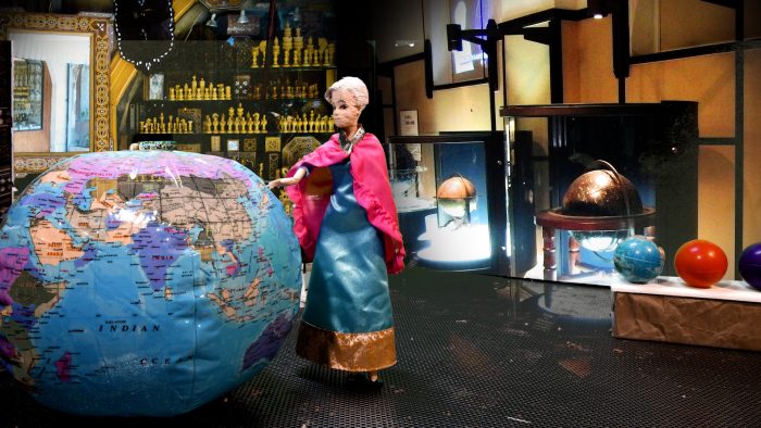 A doll in a blue dress and pink cape stands beside a large colorful globe, with smaller globes and shelves displaying chess pieces visible in the museum like background.