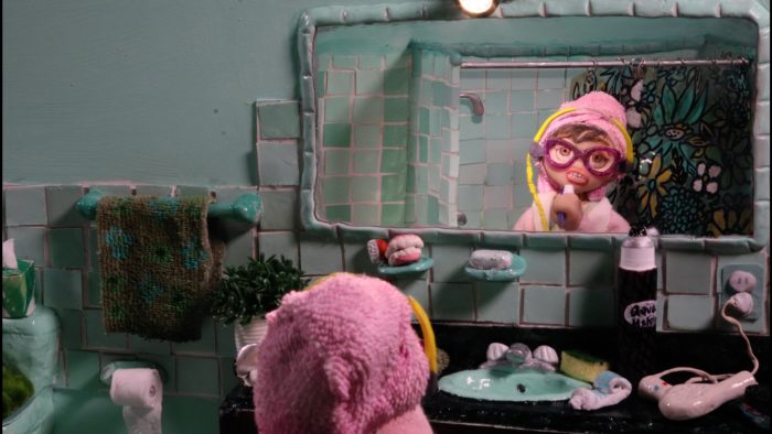 A stop motion bathroom scene features a figure with a pink towel on their head looking into a mirror, revealing another figure with glasses and a pink towel brushing their teeth.