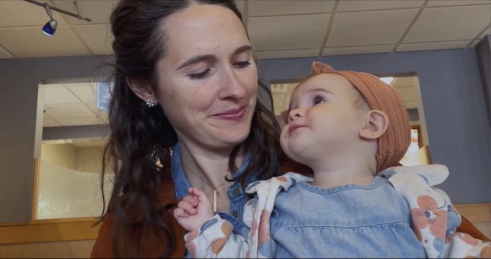 A woman with long dark hair and a gentle smile cradles a baby wearing a denim dress and an orange headband, who looks upwards in an indoor setting.