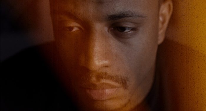 The grainy image features a close up of a mans face with a mustache, his expression pensive as one side is bathed in warm orange light and the other in shadow.