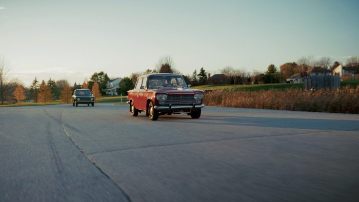 Two vintage cars, a red one in the foreground and a darker one further back, drive on a paved road winding through a residential area with houses, trees, and golden dried grass under a soft evening sky.