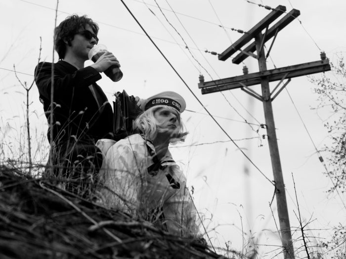 A man in sunglasses drinks from a cup as he stands behind a blonde person in a CHOO CHOO sailor hat, both looking out from a grassy foreground with a utility pole in the background.