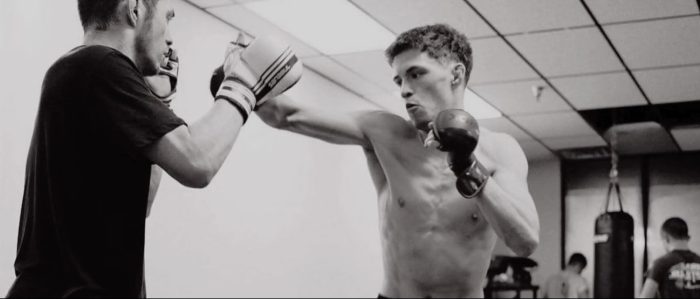 A black and white image captures two men boxing or training in a gym, with one shirtless man throwing a punch and another man in a dark shirt holding a block.