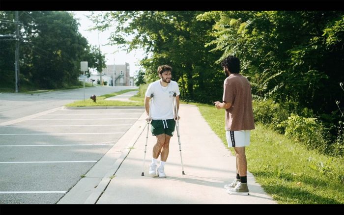 Two men are conversing on a sunny sidewalk, one walking with crutches and the other standing, with a street and trees in the background.