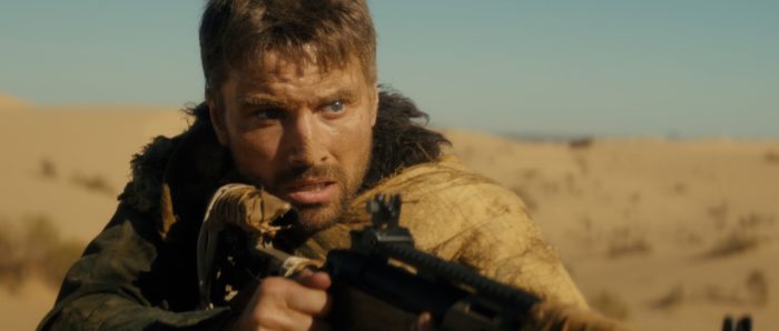A man with a serious expression holds a rifle in a barren desert environment under a clear blue sky.