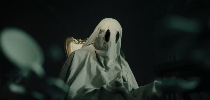 A figure resembling a ghost, covered in a white sheet with dark eyeholes, sits on an ornate golden chair amidst blurred dark surroundings.