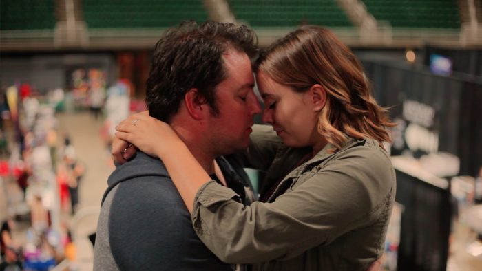 A man and woman embrace with their foreheads touching and eyes closed, standing within a large, out of focus indoor environment.