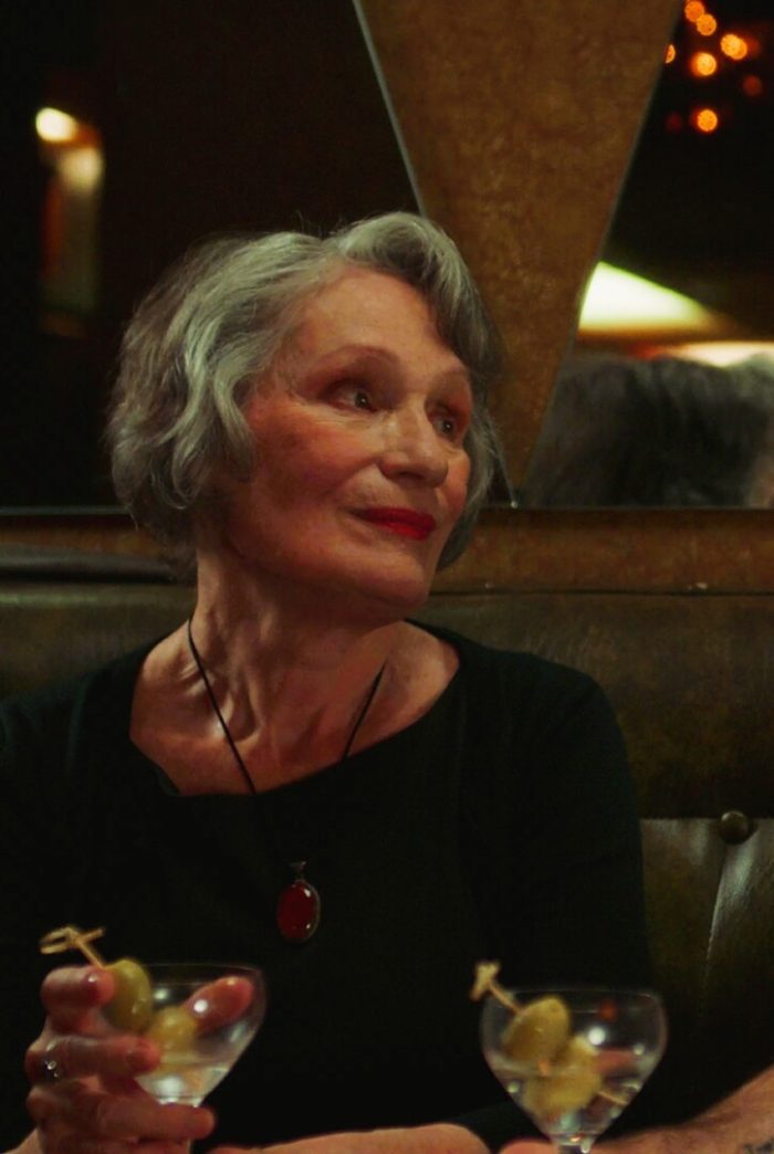 An older woman with gray hair and red lipstick holds a martini glass containing olives, wearing a red pendant necklace and looking to her right in a dimly lit setting.