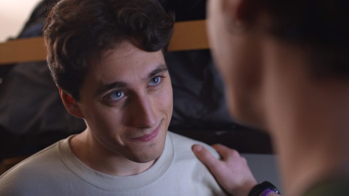 A young man with curly brown hair and blue eyes smiles warmly while looking up, as another persons hand gently rests on his shoulder.