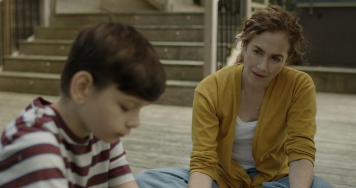 A woman wearing a yellow cardigan and blue jeans sits on a wooden deck, gazing towards a young boy in a striped shirt who is looking down in the blurry foreground, with stairs visible behind them.