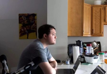 A man in a gray shirt sits in profile at a counter with two laptops and a microphone, looking right towards a kitchen area filled with various appliances and wooden cabinets, with a movie poster hanging on the wall to his left.