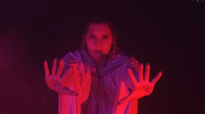 A woman wearing a shiny hooded garment extends both hands forward with a direct gaze, illuminated by vibrant red and purple light against a dark, smoky backdrop.