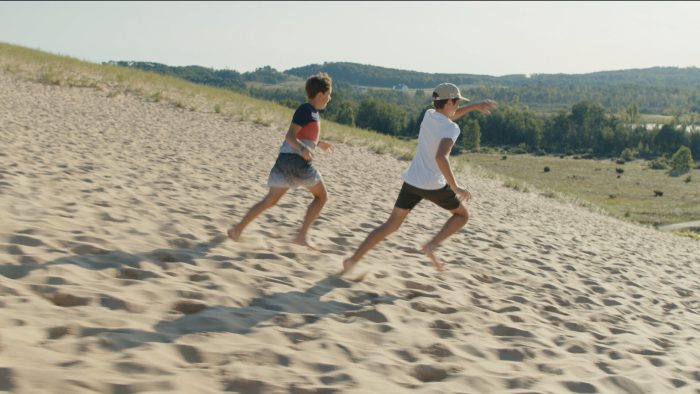 Two boys run barefoot down a large sand dune under a bright sky, with a forested landscape and distant water in the background.