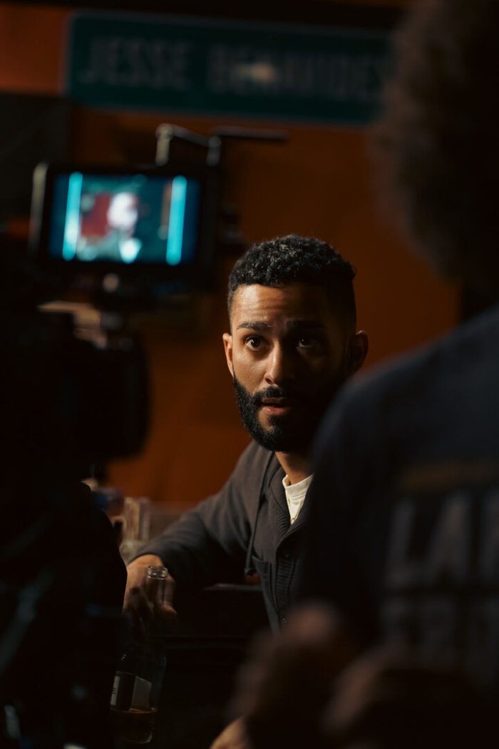 A bearded man in a dark cardigan looks intently forward from a bar setting, with another person blurred in the foreground and a monitor and sign visible in the background.