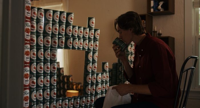 A young man sits amidst an elaborate structure made of stacked tomato soup cans, intently smelling a single can.
